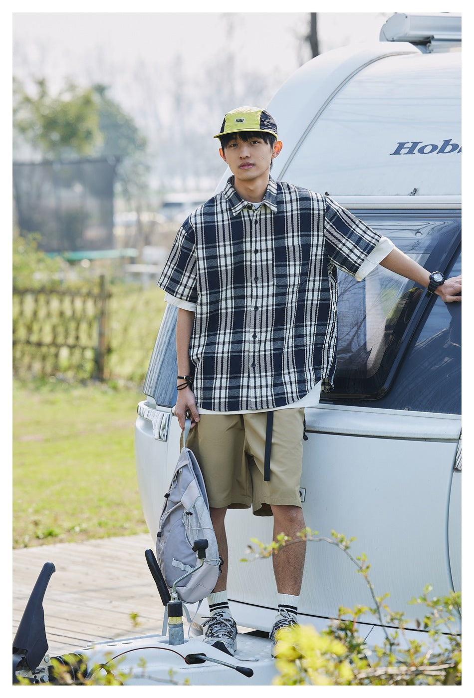 Model wearing a navy and white plaid short-sleeve shirt with khaki shorts, posing beside a vehicle.