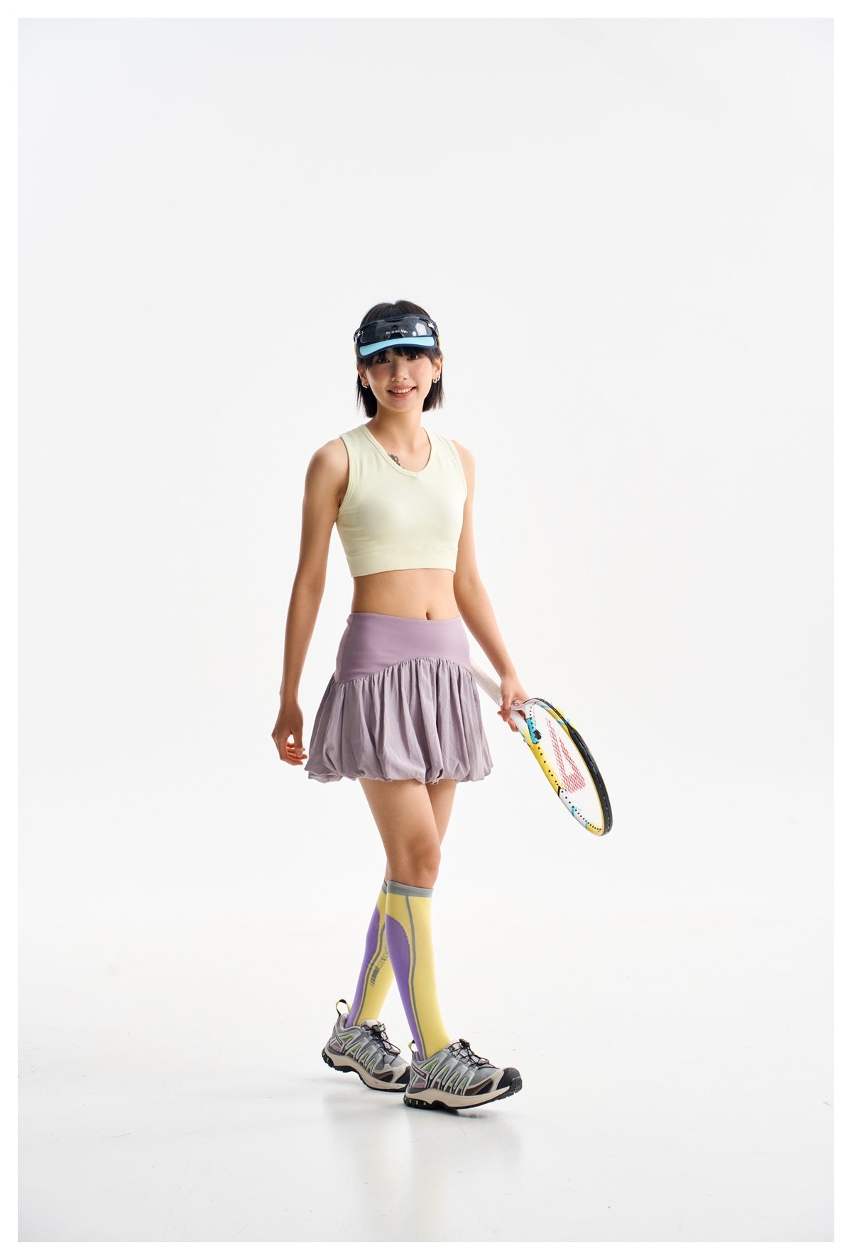 Model wearing a pale yellow crop tank and lilac bubble-hem skirt, holding a racket against a white studio background.