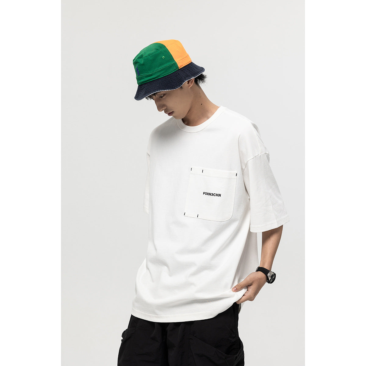 Model wearing an oversized white T-shirt with a stitched chest pocket and black text, paired with black shorts.