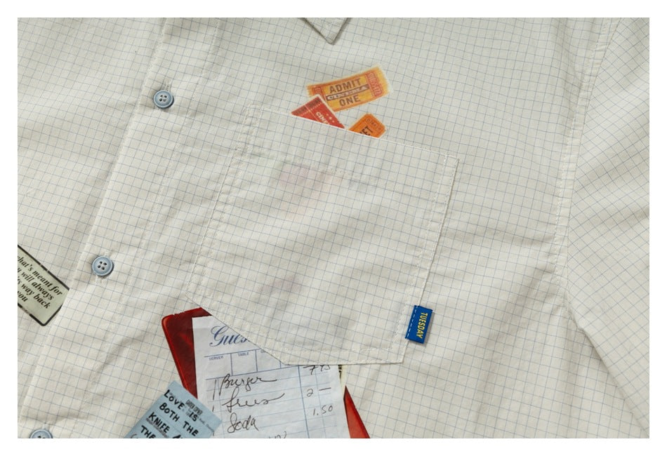 Close-up of a cream blue-grid shirt chest pocket, buttons, and colorful ticket and paper prints.