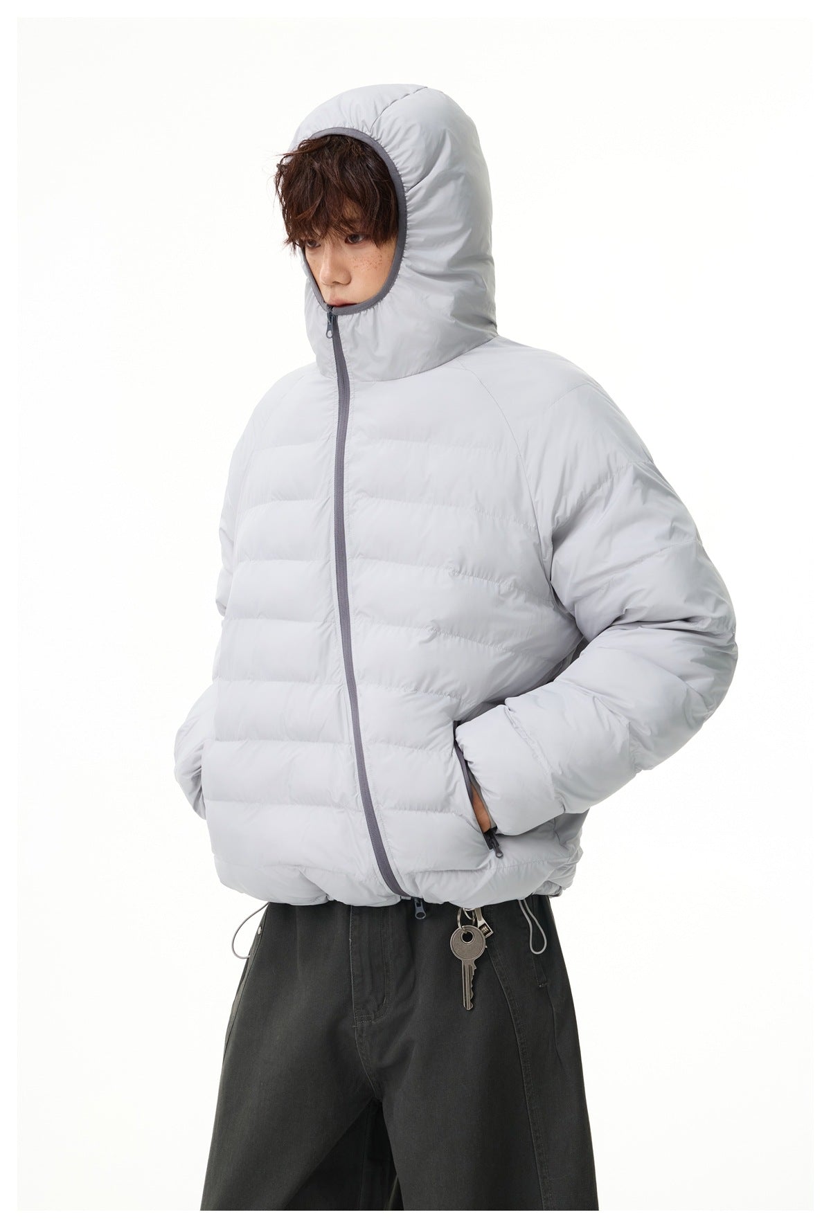 Model wearing a light gray hooded quilted jacket with the hood up, hands in pockets, with dark pants and keys