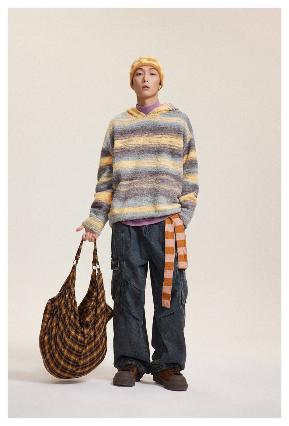Model wearing the gray and yellow striped hooded knit sweater with wide jeans, holding a plaid bag