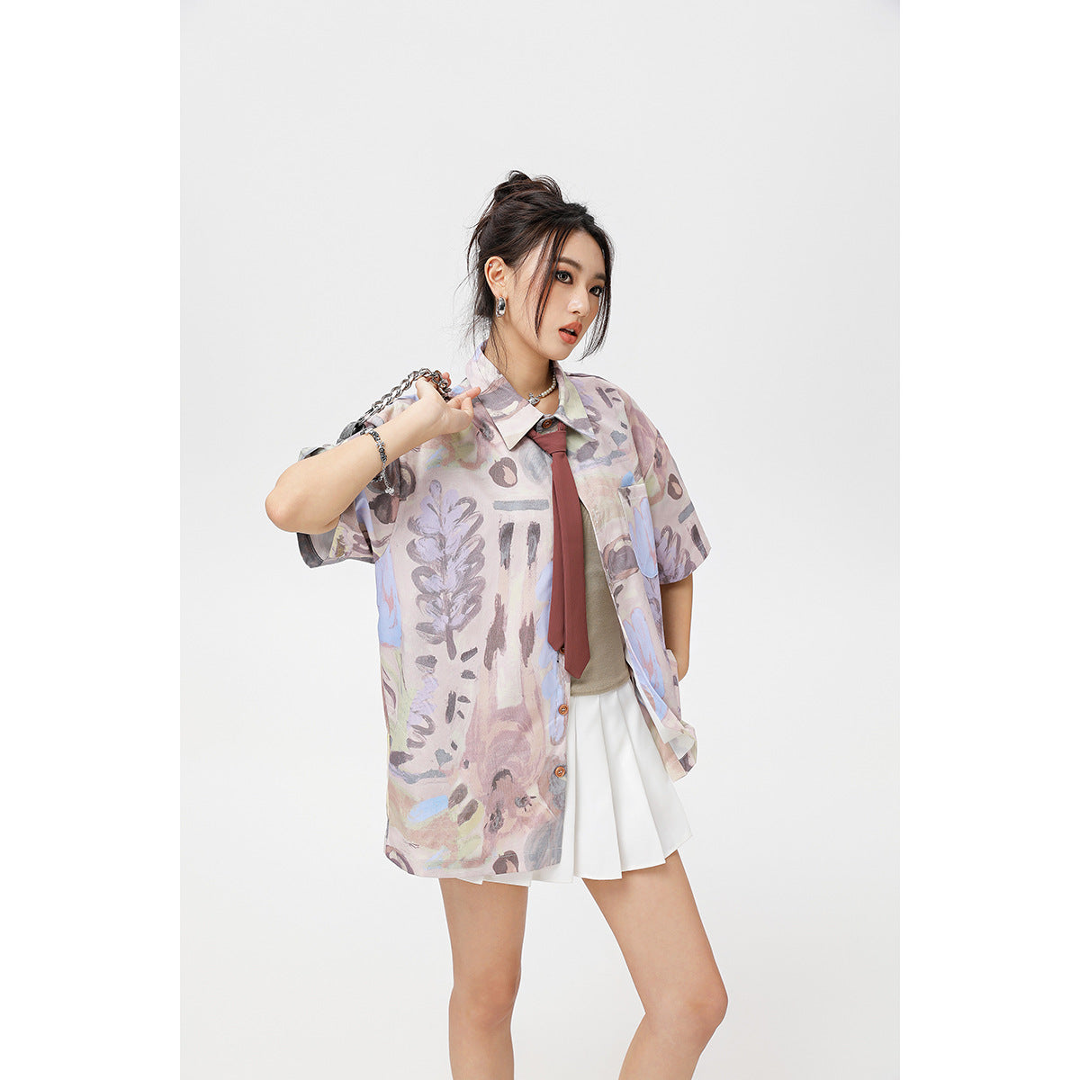 Three-quarter view of a model wearing a pastel abstract-print oversized shirt with a burgundy tie and white skirt.