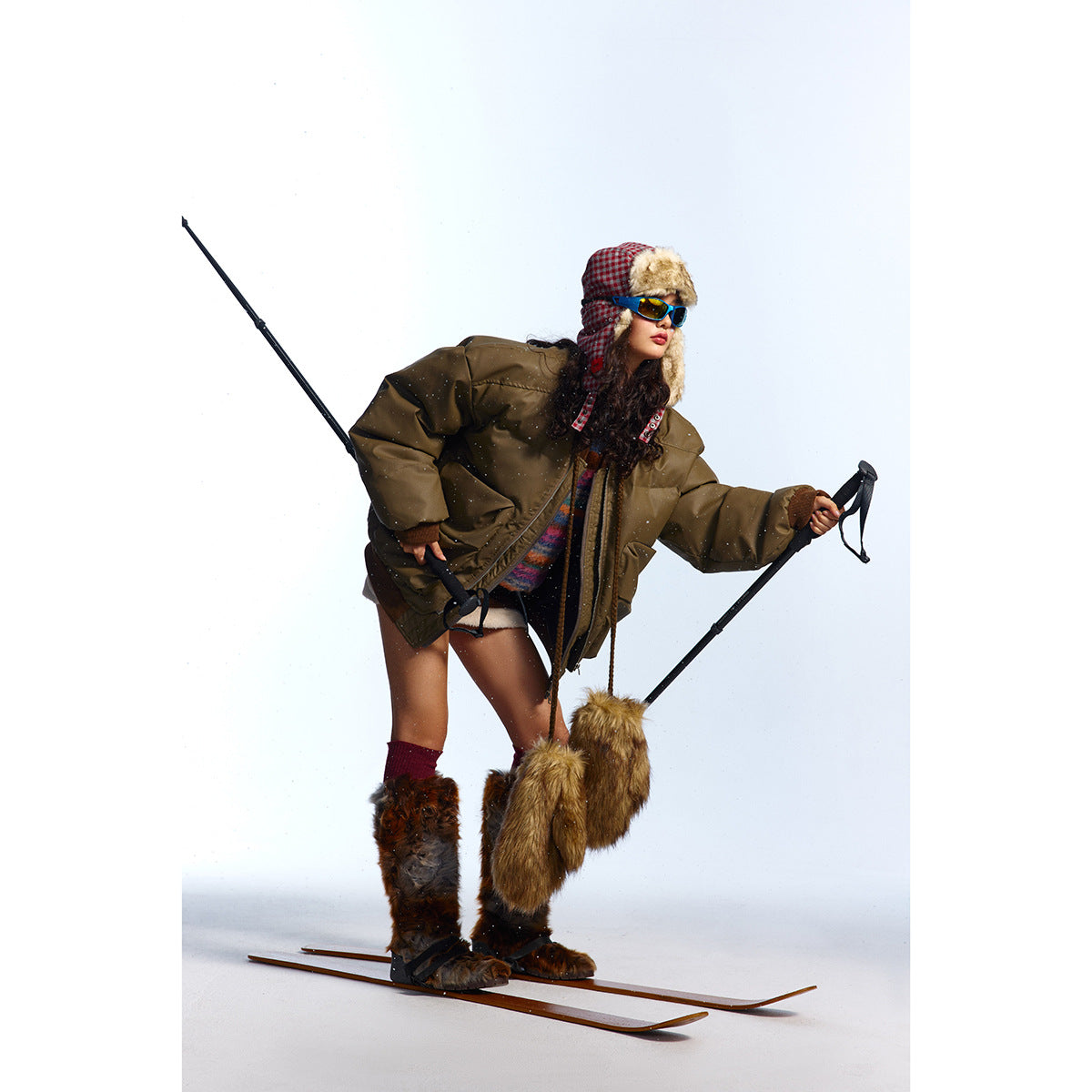Model wearing an oversized olive-brown puffer jacket with a striped sweater and winter accessories, posed on skis.