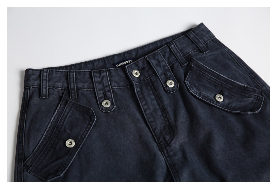 Close-up of dark charcoal jeans showing the waistband, belt loops, metal-button tabs, and flap pockets.