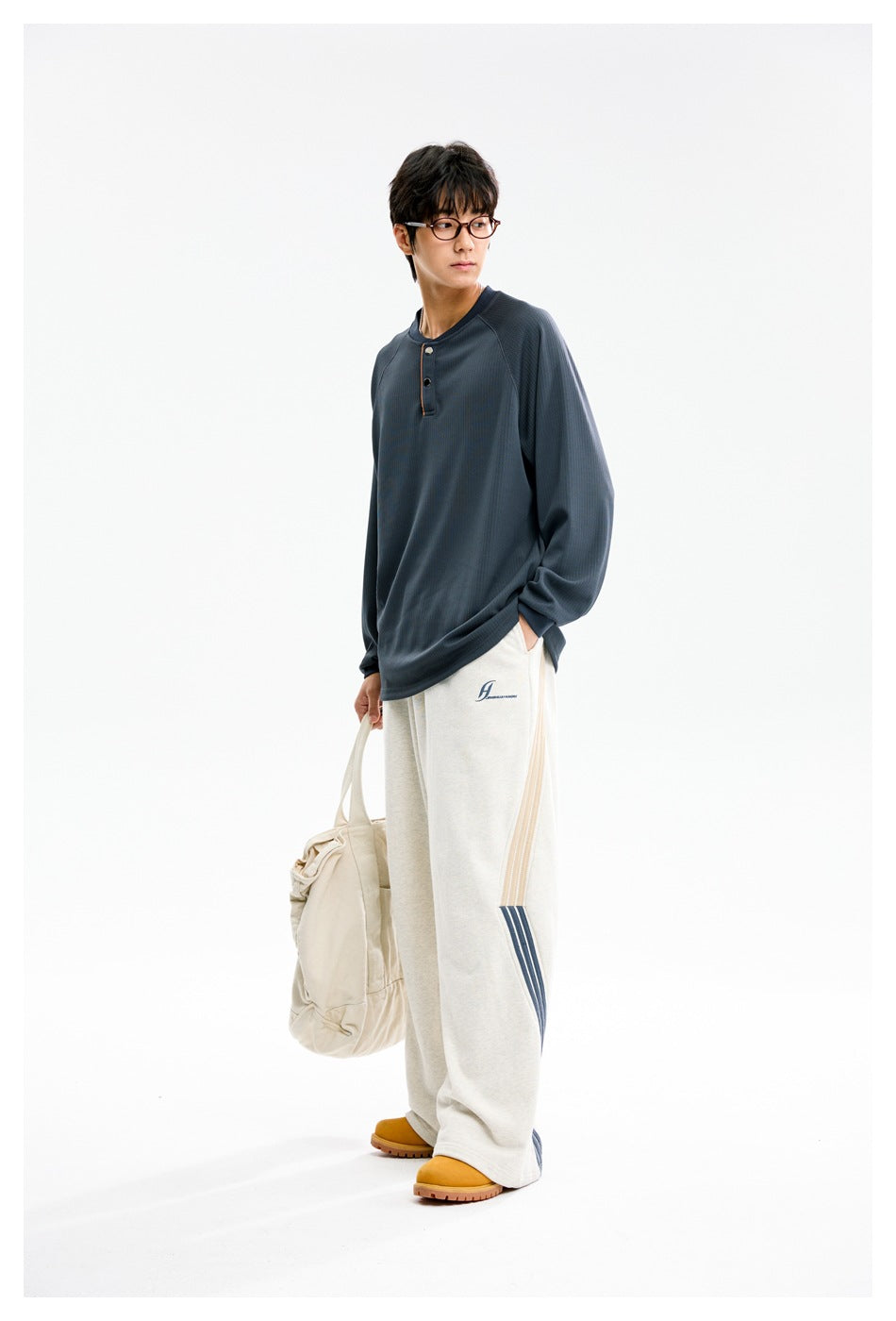 Model in a blue ribbed henley top, angled view, with off-white striped wide-leg pants and a cream tote