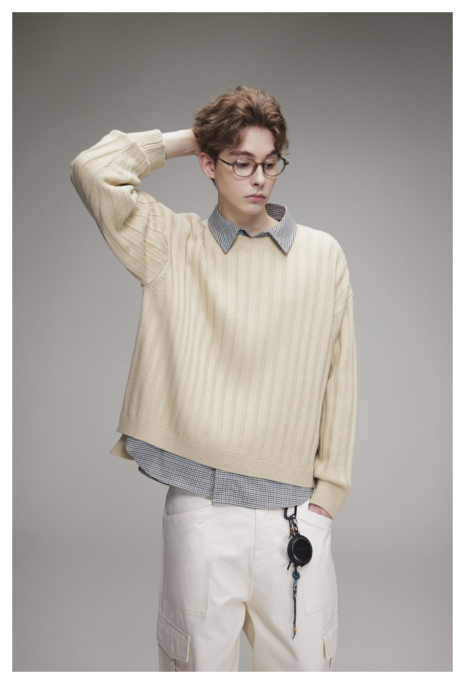 Model wearing a cream wide-rib knit sweater over a checked collared shirt with off-white cargo pants, hand behind head