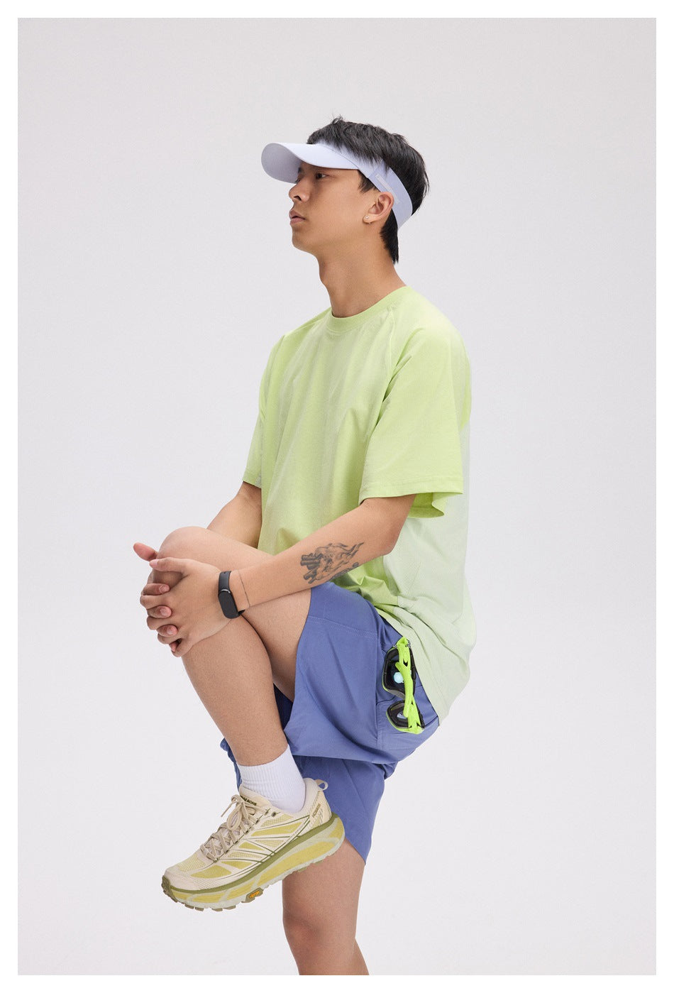 A seated man wears a pale lime short-sleeve T-shirt and blue-violet shorts, shown from the side.