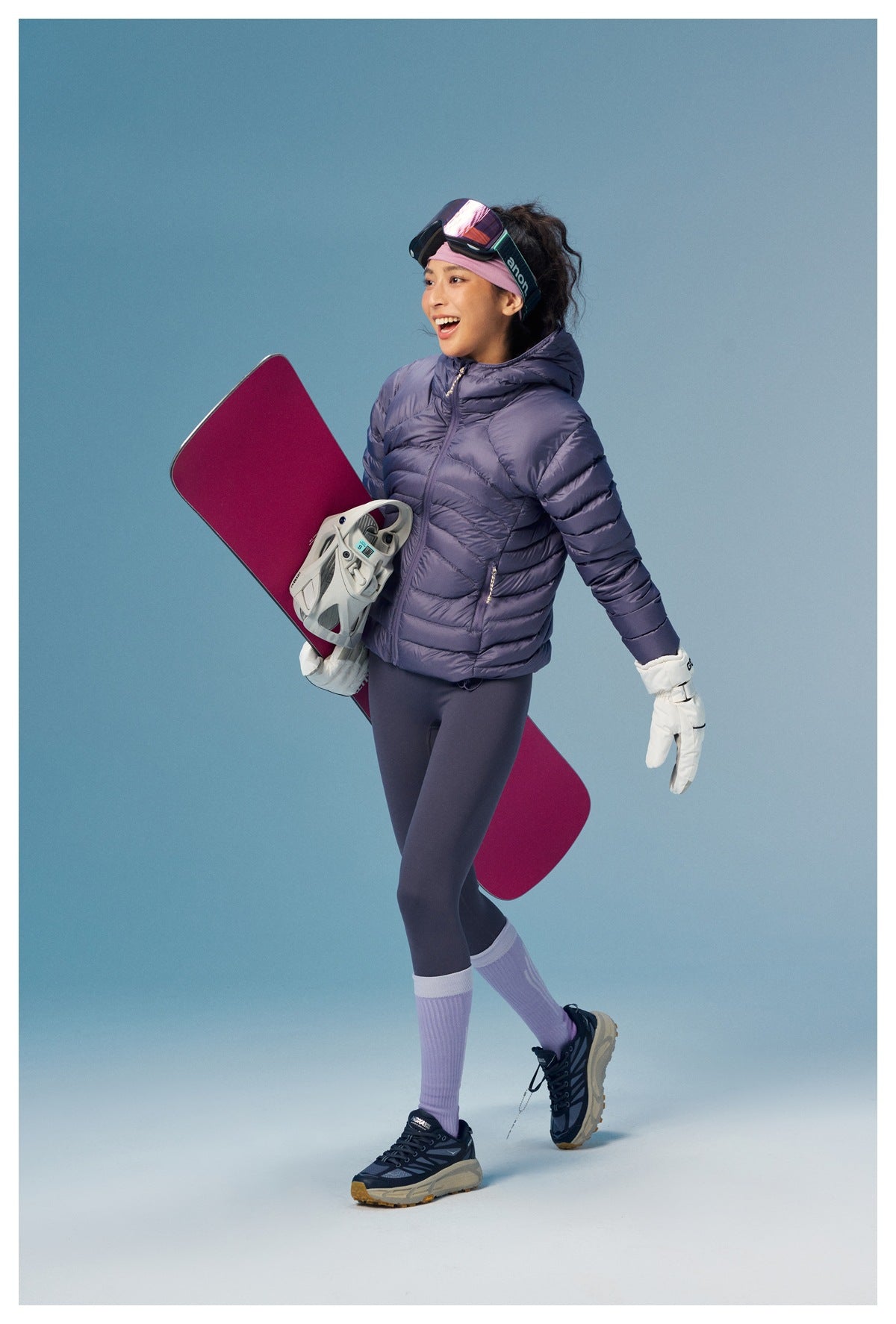 Woman walking in a purple hooded quilted puffer jacket and leggings, carrying a snowboard, side view