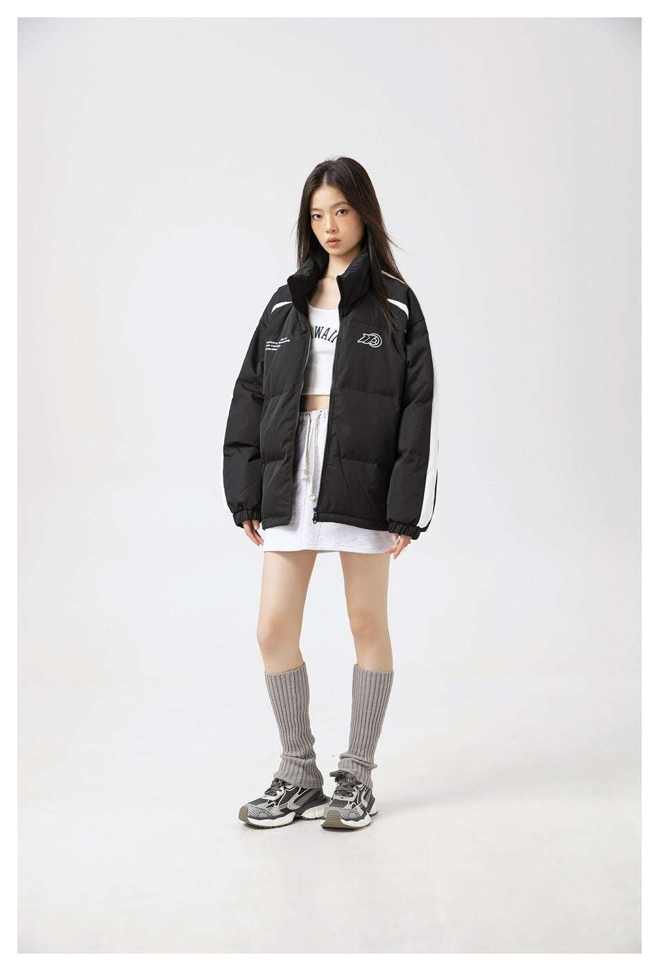 Model wearing a black puffer jacket with white sleeve stripes, open over a white tank top and gray skirt, front view