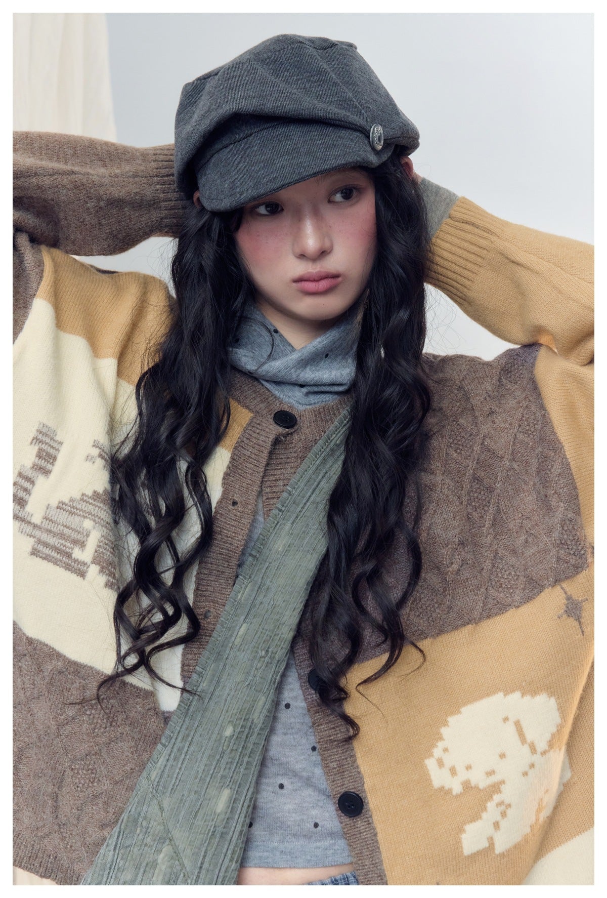 Close-up of a model wearing an oversized brown, mustard and cream color-block cardigan with graphic motifs and a gray cap.