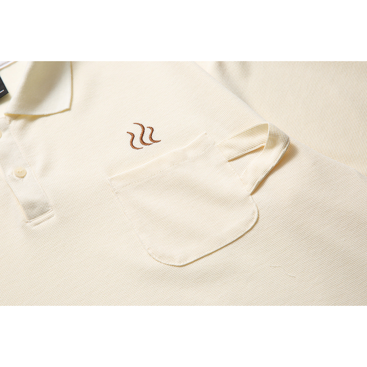 Close-up of an ivory polo shirt's chest pocket, short sleeve, and small brown embroidered motif.