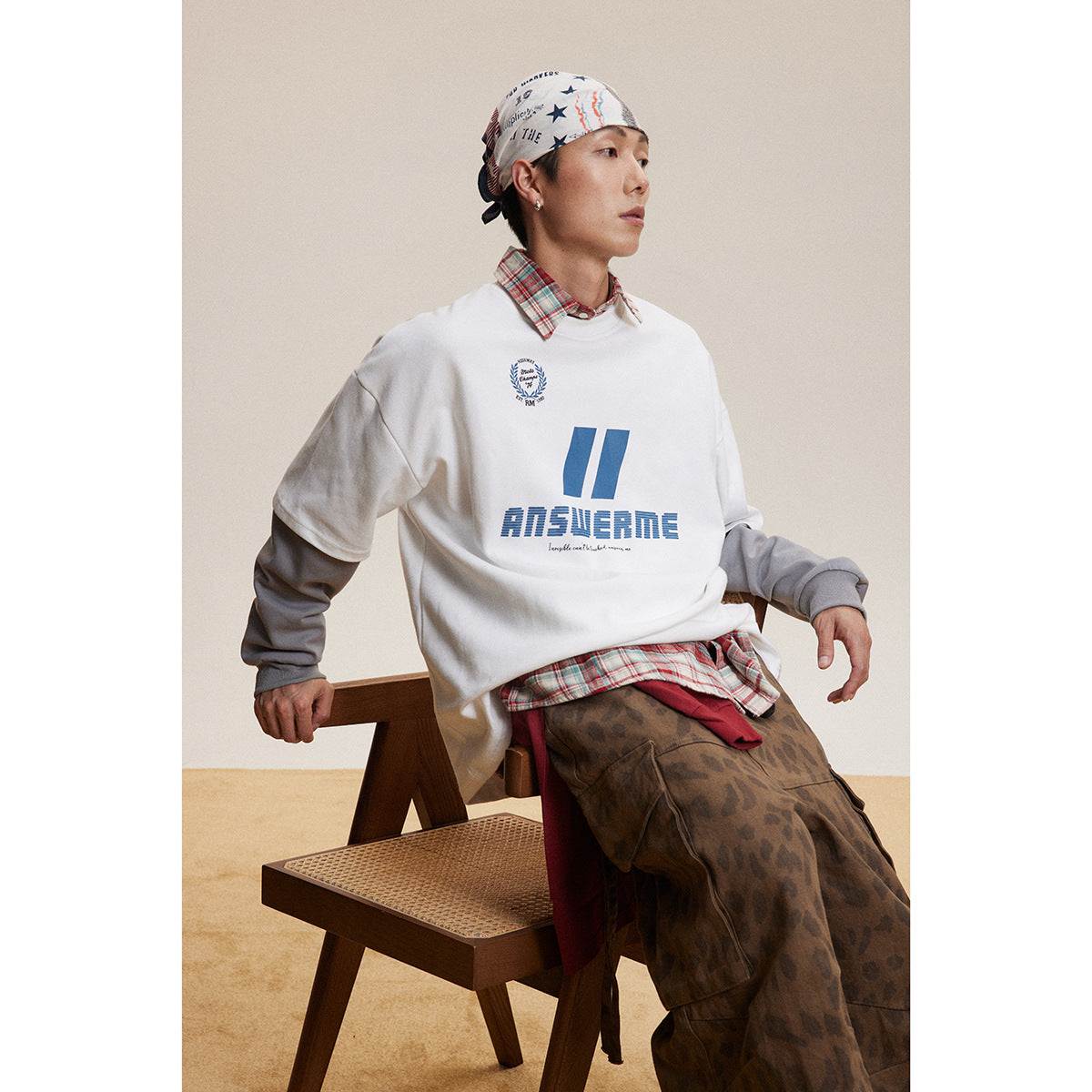 Model seated in a white oversized sweatshirt with blue chest graphics, layered over a plaid shirt and gray sleeves.