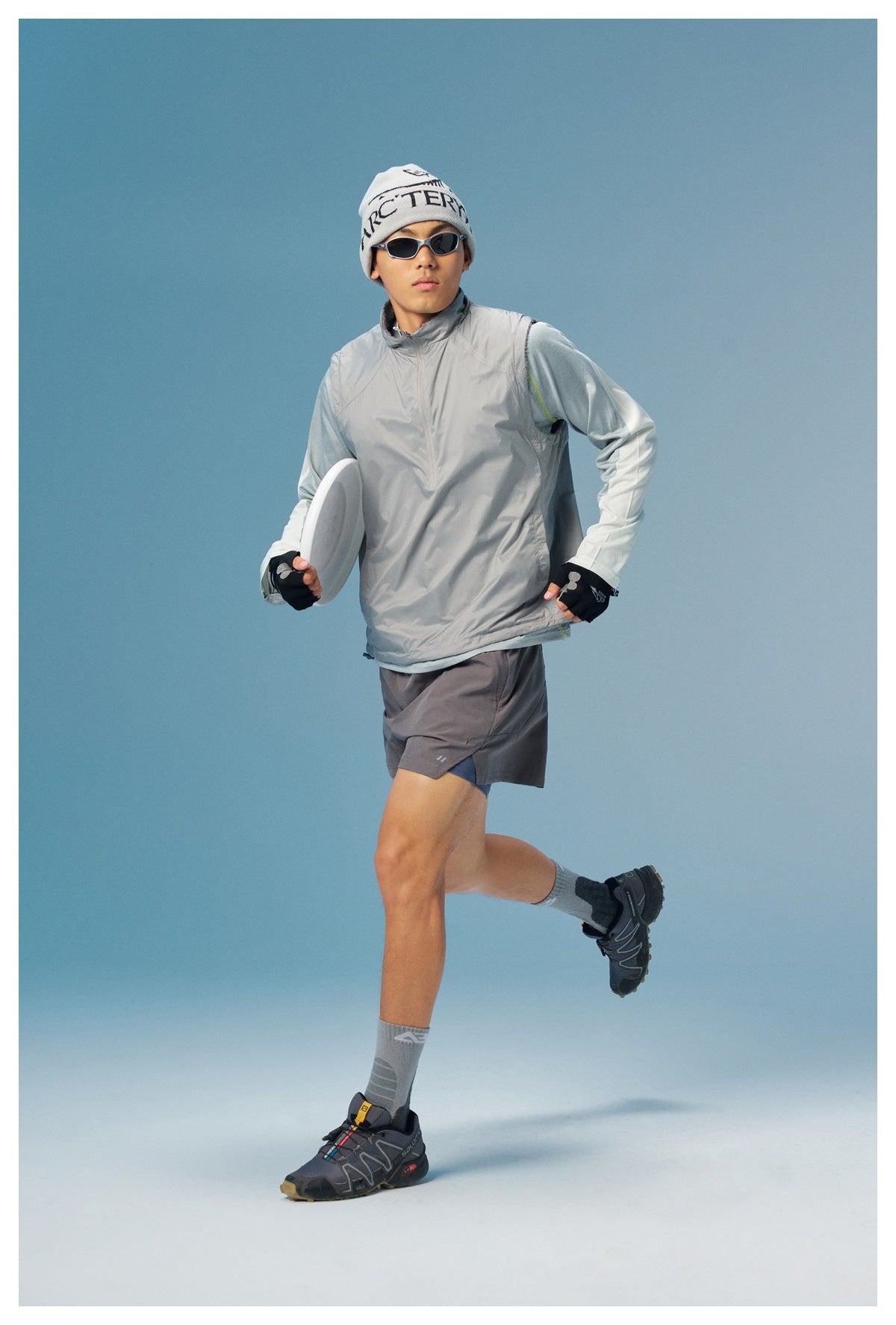 Man modeling a light gray sleeveless zip-neck vest over a pale long-sleeve top, with gray shorts.