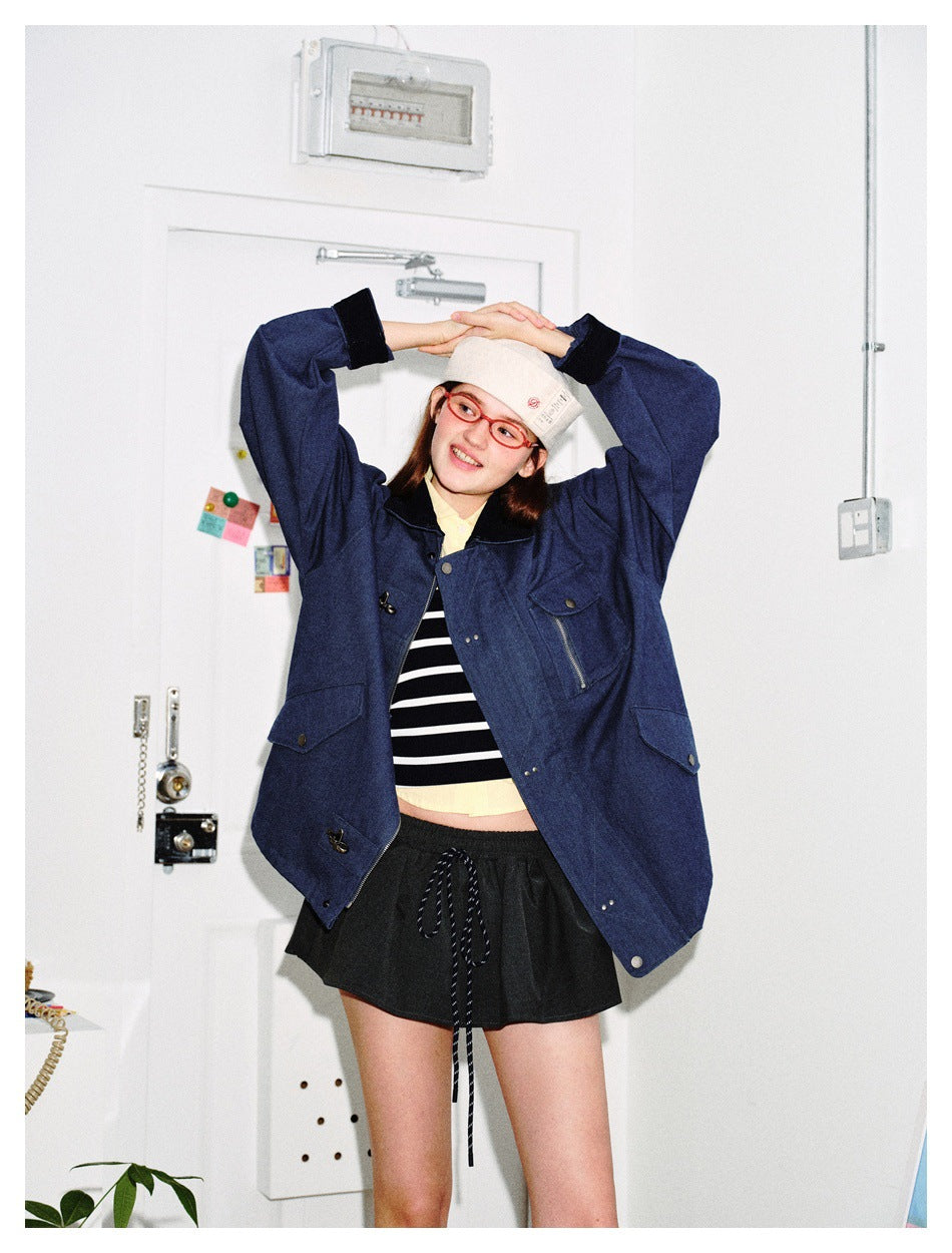 Model wearing an open navy jacket over a striped top and black shorts, arms raised over a cap in a white room