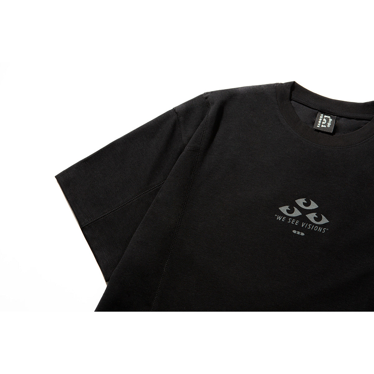 Close-up of a black crew-neck T-shirt showing its short sleeve, seams, neck label, and small eye graphic with “WE SEE