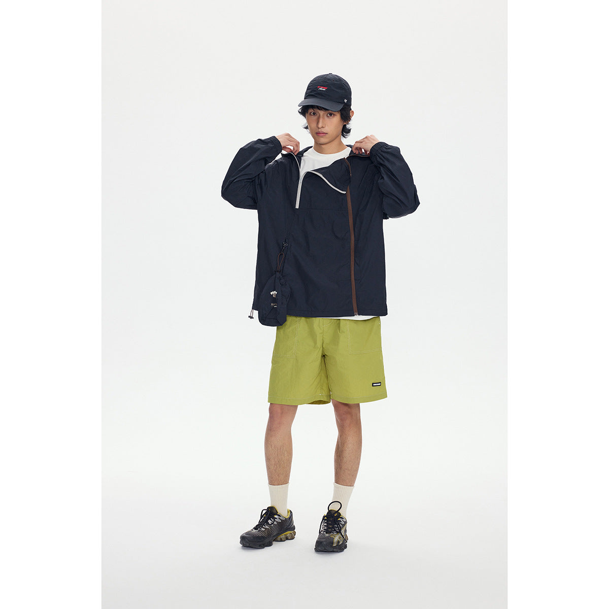 Model wearing a dark hooded pullover jacket with contrasting zips and lime-green shorts, shown from the front.