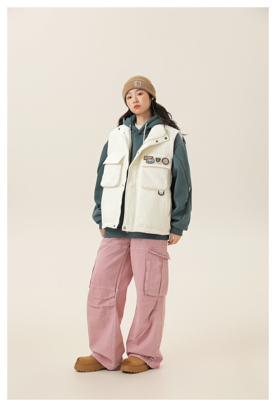 Model wearing off-white textured vest over a teal hoodie with pink cargo pants and tan boots, full-length view