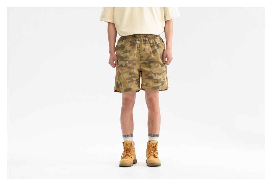 Person wearing tan and brown camouflage shorts with a cream tee and yellow work boots, front view cropped at the torso