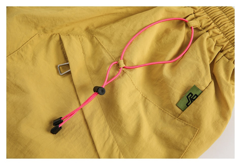 Close-up of mustard shorts showing a front pocket, pink drawcord and toggle, and a small woven tag.