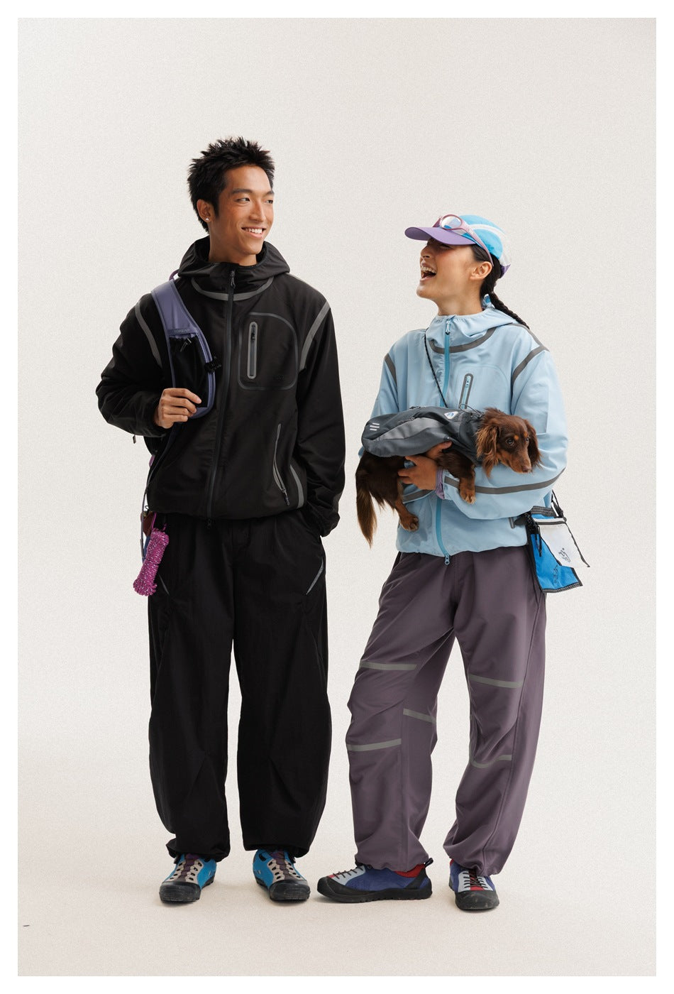 Two models wear outdoor jackets and loose technical pants; one holds a small dog.