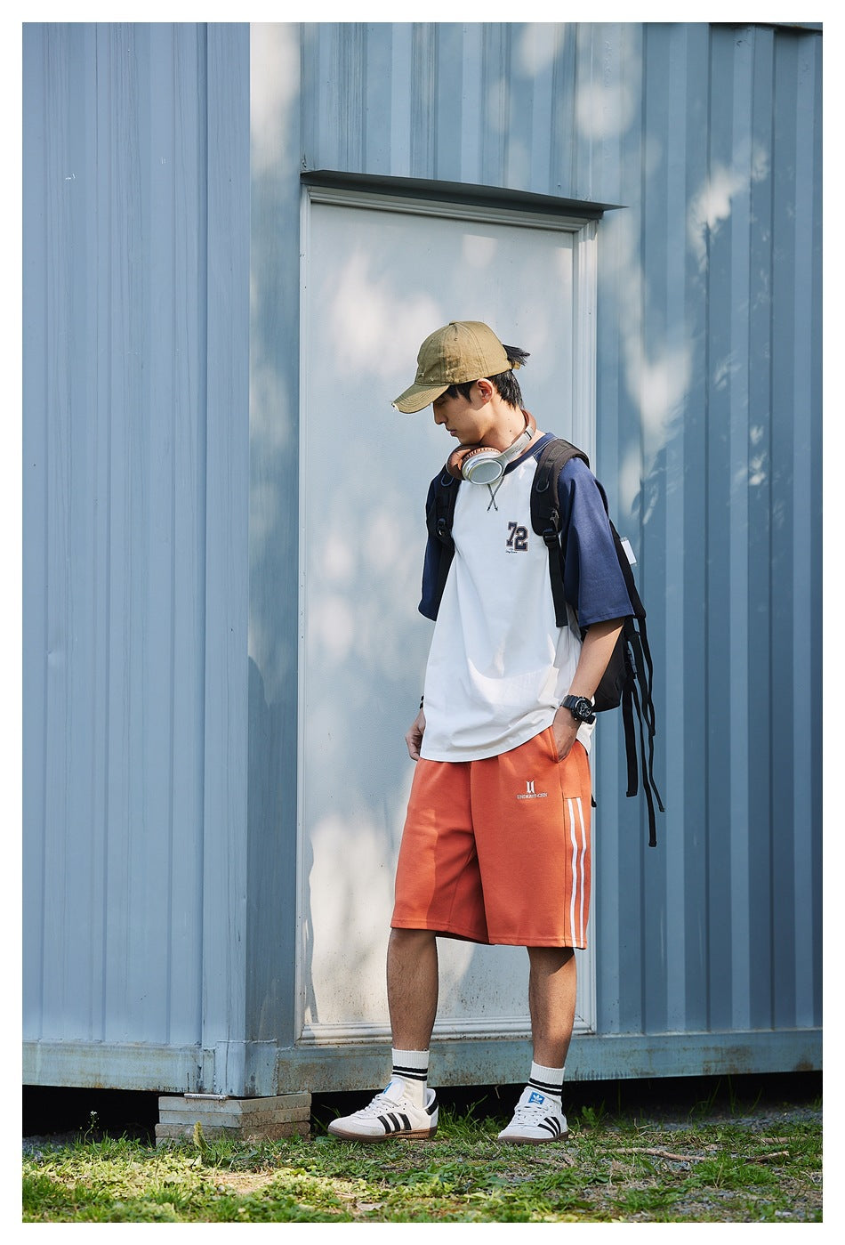 Model wearing the white T-shirt with navy raglan sleeves and 72 chest print, with orange shorts, cap and backpack