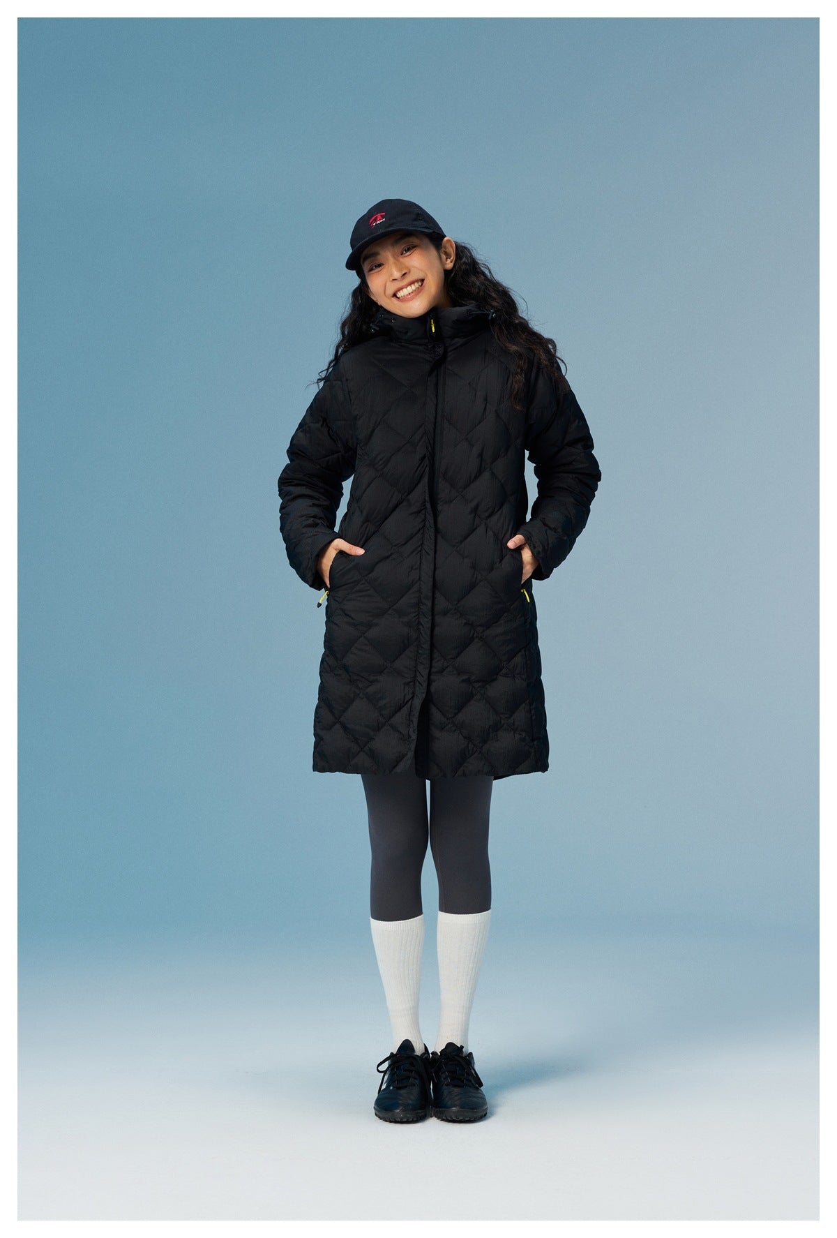 Smiling woman wearing a black long quilted hooded jacket zipped up with hands in the pockets, cap and white socks
