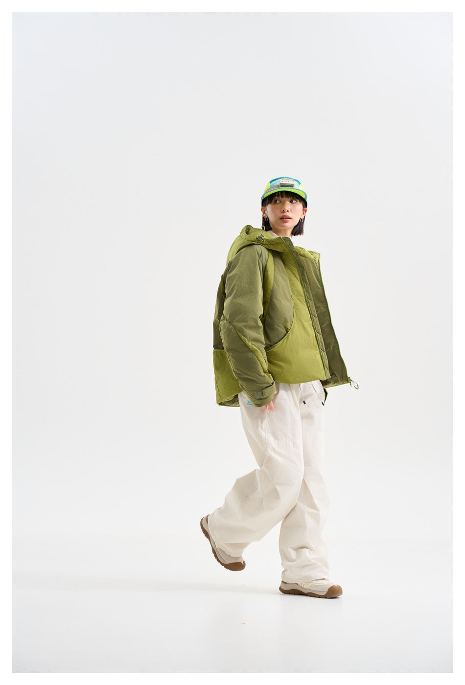 Model walking in a side view of an olive and lime-green color-blocked hooded jacket with loose white trousers.