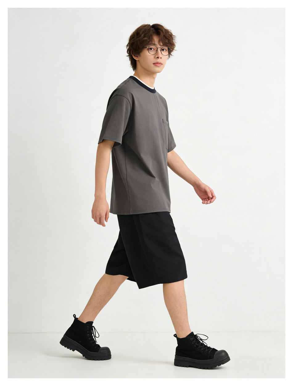 Model walking in a dark gray pocket T-shirt with navy collar, black shorts and black boots, side view