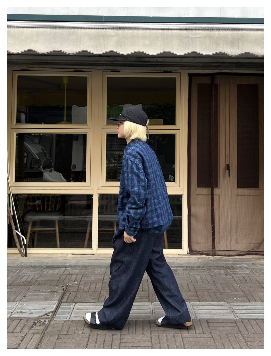 Person wearing a blue plaid shirt, loose dark trousers, and a black cap outdoors, shown in profile.