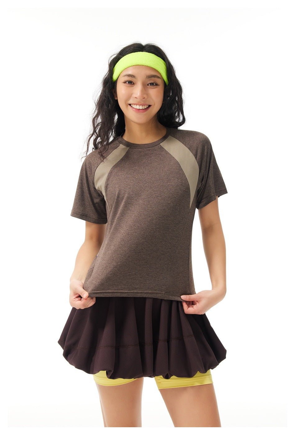 Model wearing a brown short-sleeve athletic top with taupe panels, a dark flared skirt, yellow shorts and a neon headband.