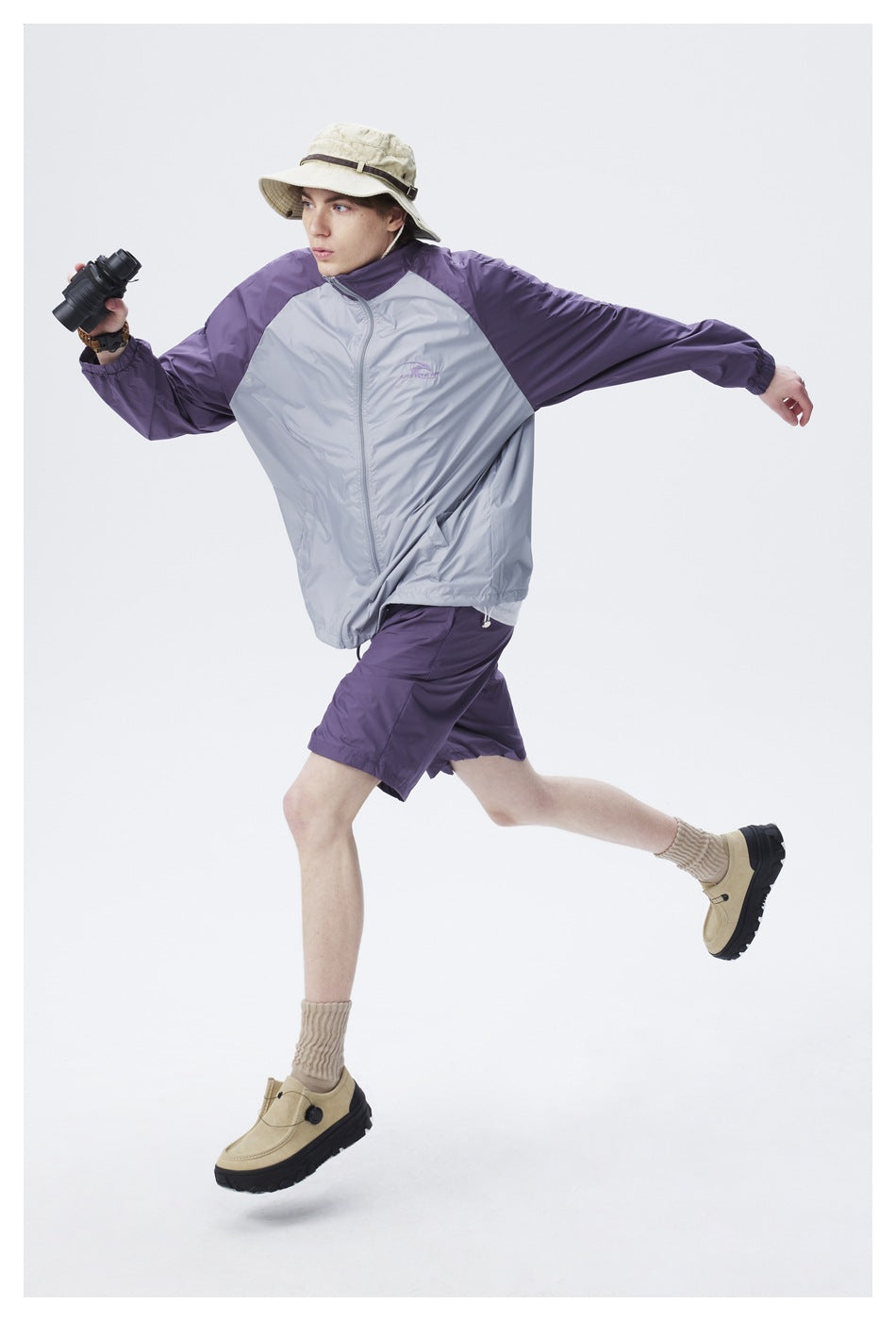 Model in a gray windbreaker with purple sleeves and purple shorts, wearing a beige bucket hat and holding binoculars.