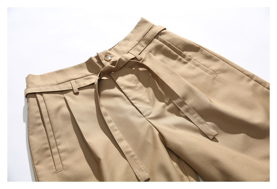 Flat-lay view of tan trousers with a button waist, pleats, belt loops, and a long matching tie belt.