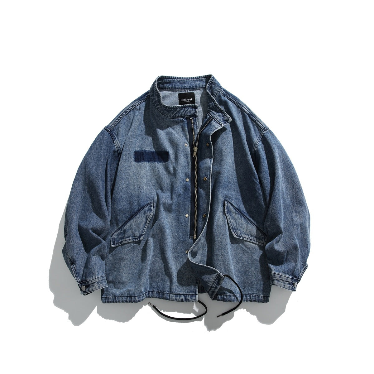 Front view of a washed blue denim jacket with a stand collar, snap-front placket, two angled pockets and black hem drawcords.