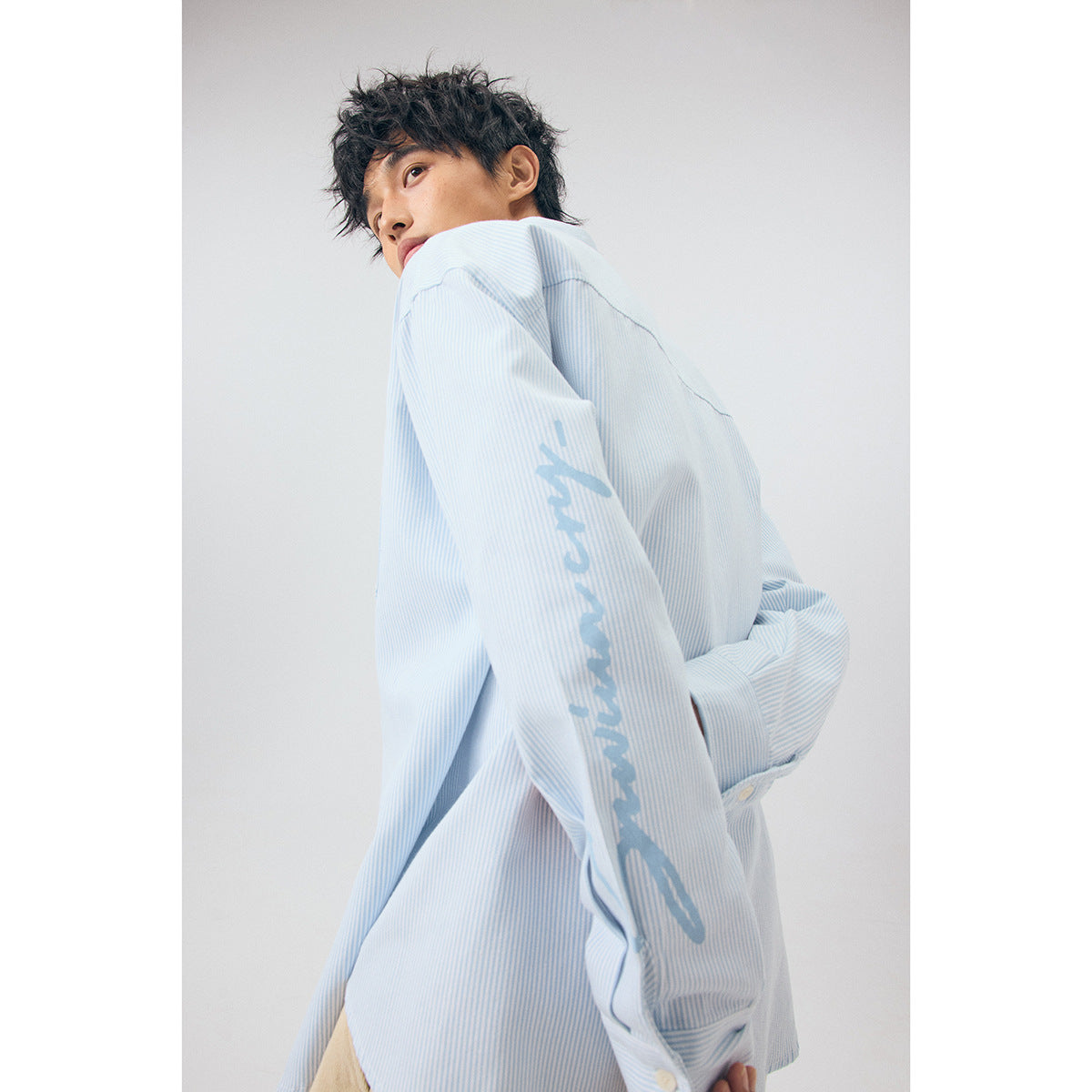 Close side view of a model wearing a pale blue pinstriped shirt with script lettering on the sleeve.