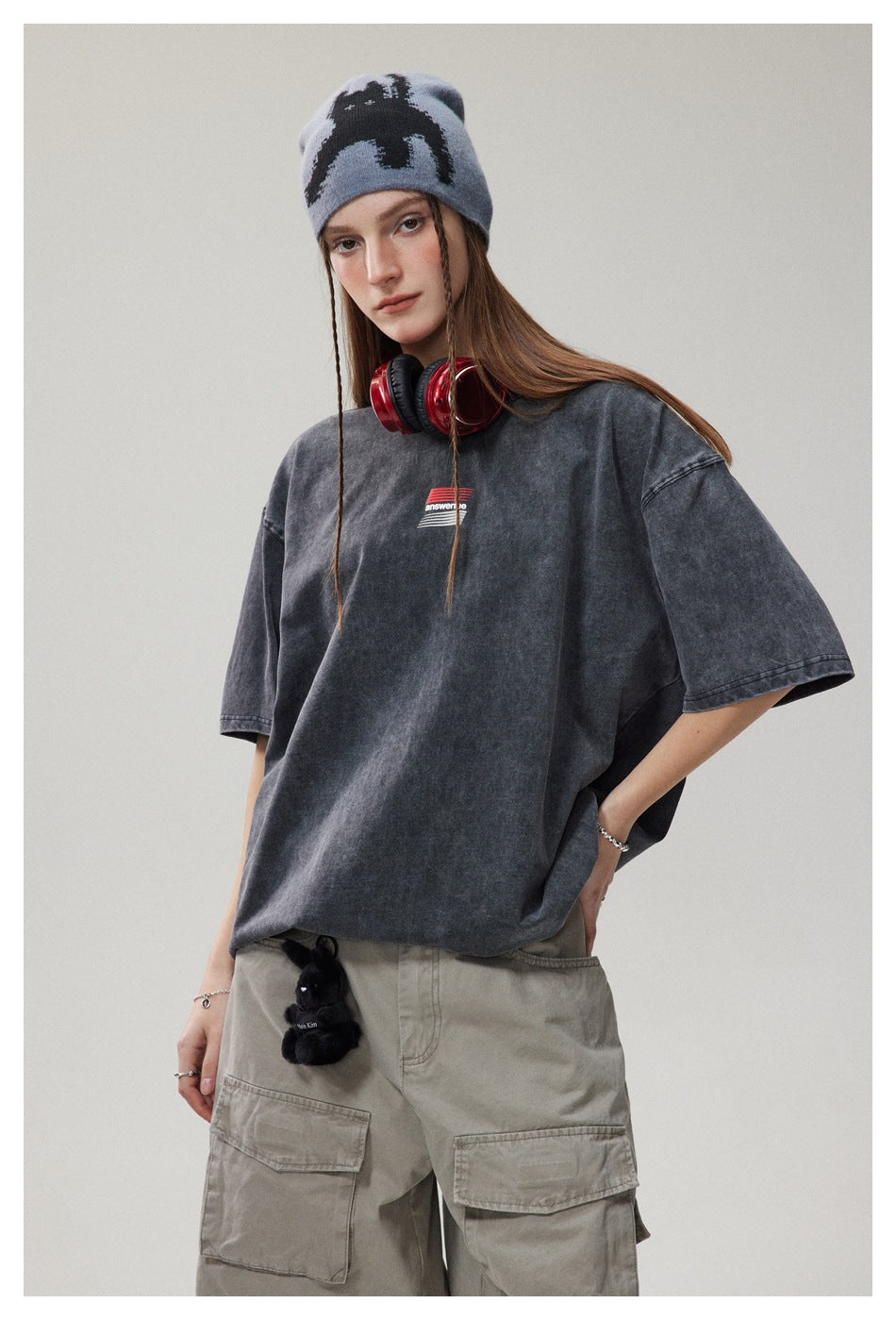 Model wearing an oversized charcoal washed T-shirt with a small red-and-white chest graphic and a patterned blue-gray beanie.