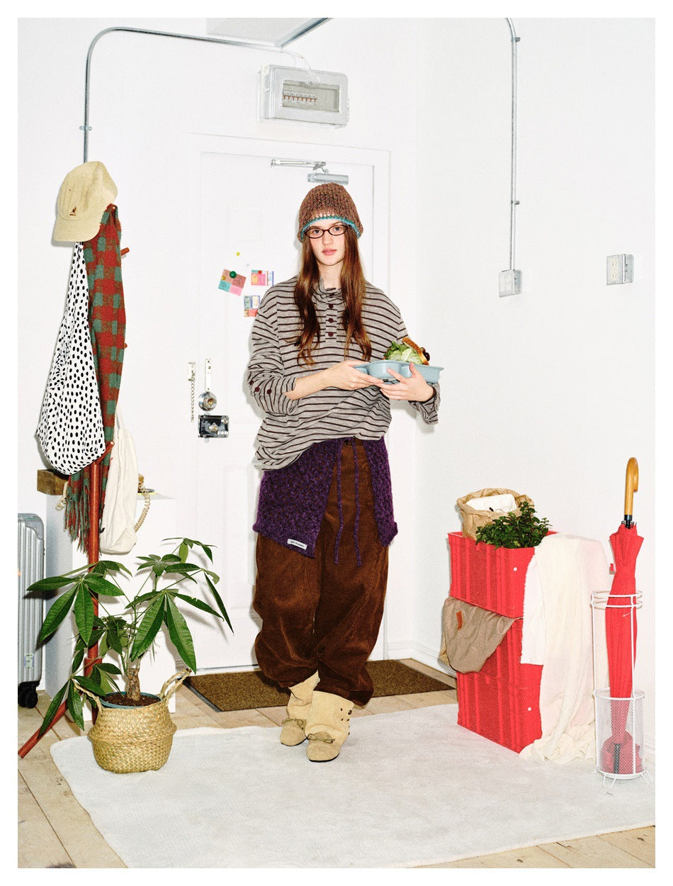 Model wearing a gray striped long-sleeve top with button details, brown trousers, a purple waist wrap, and a knit beanie.