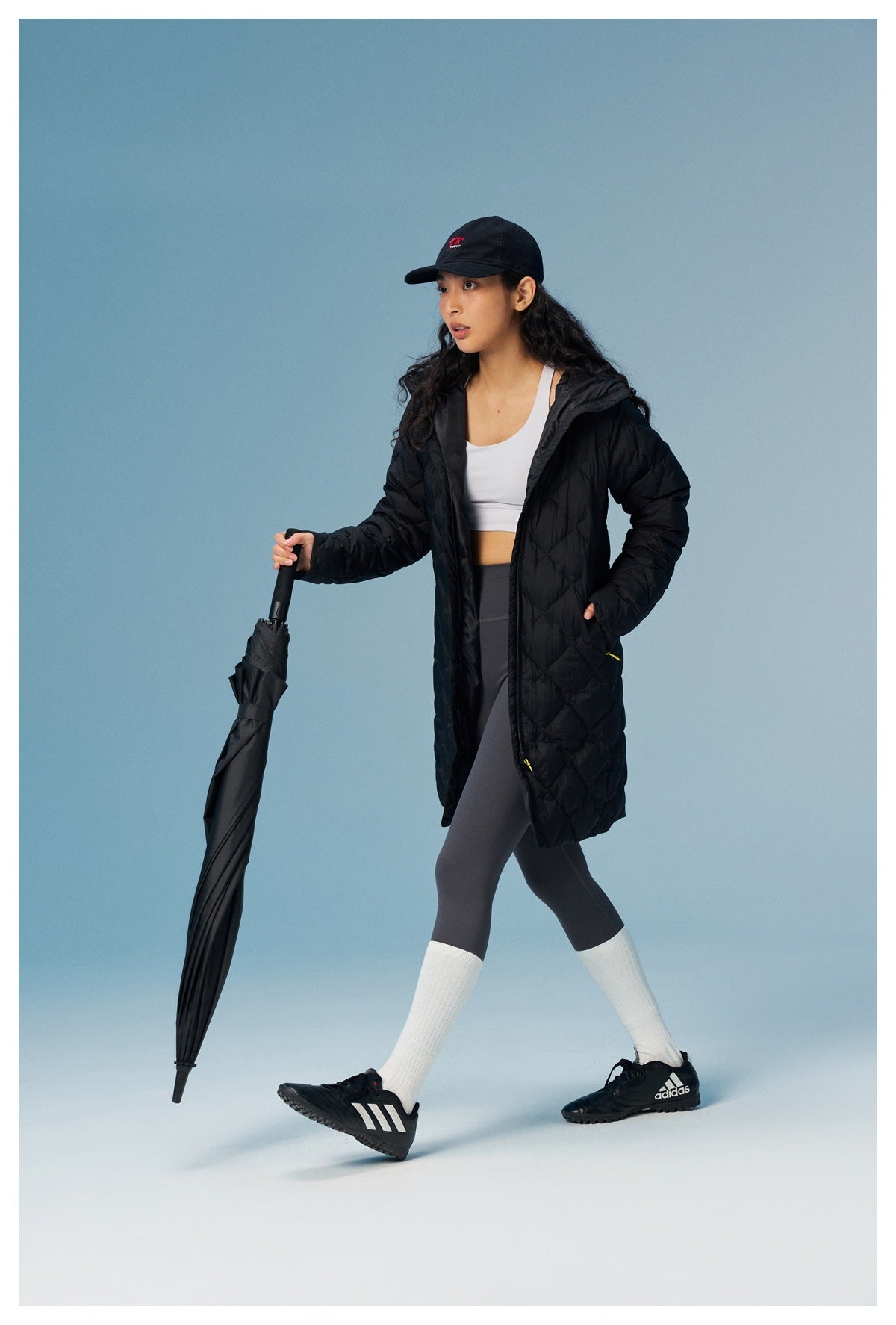 Woman walking in a black long quilted hooded jacket open over a white top and gray leggings, holding a closed umbrella