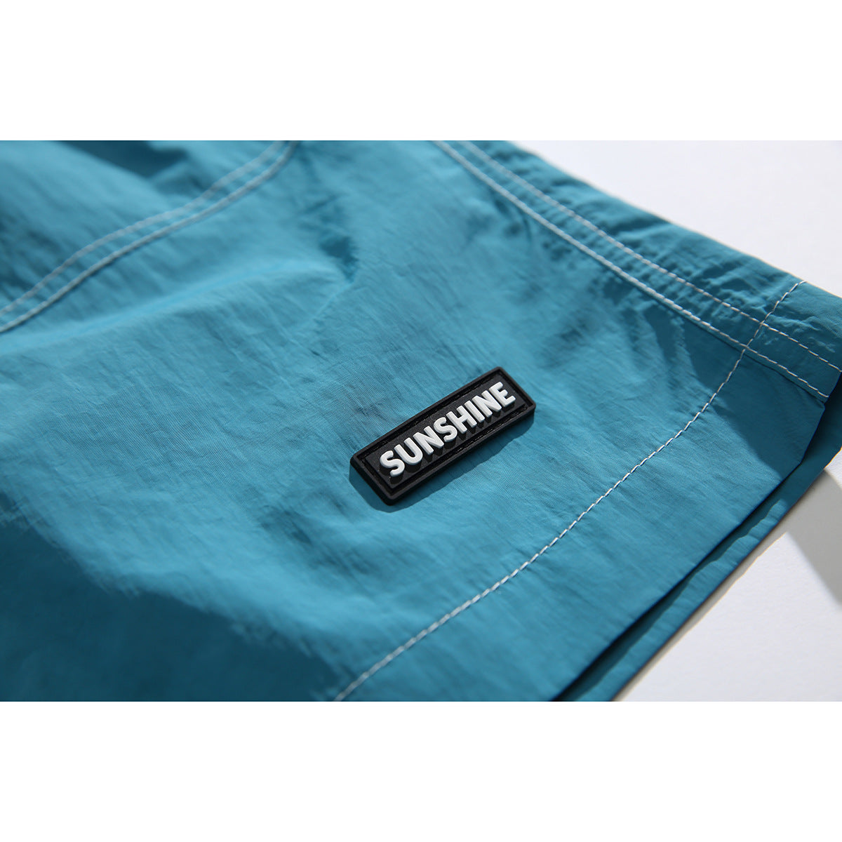 Close-up of a black SUNSHINE patch and white stitching on teal shorts.