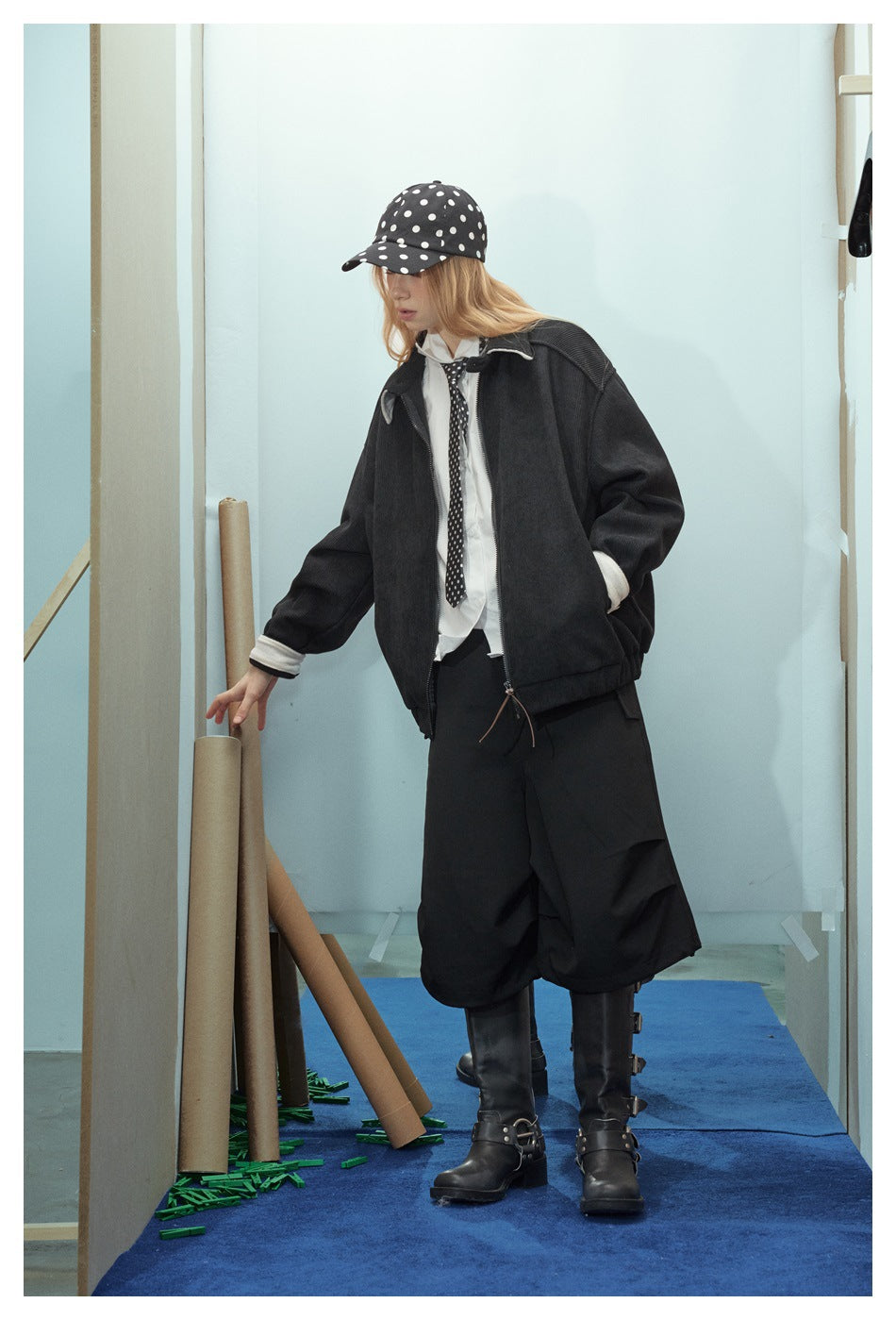 Model wearing an oversized black jacket with cream trim, shown at an angle with a polka-dot cap and tie.
