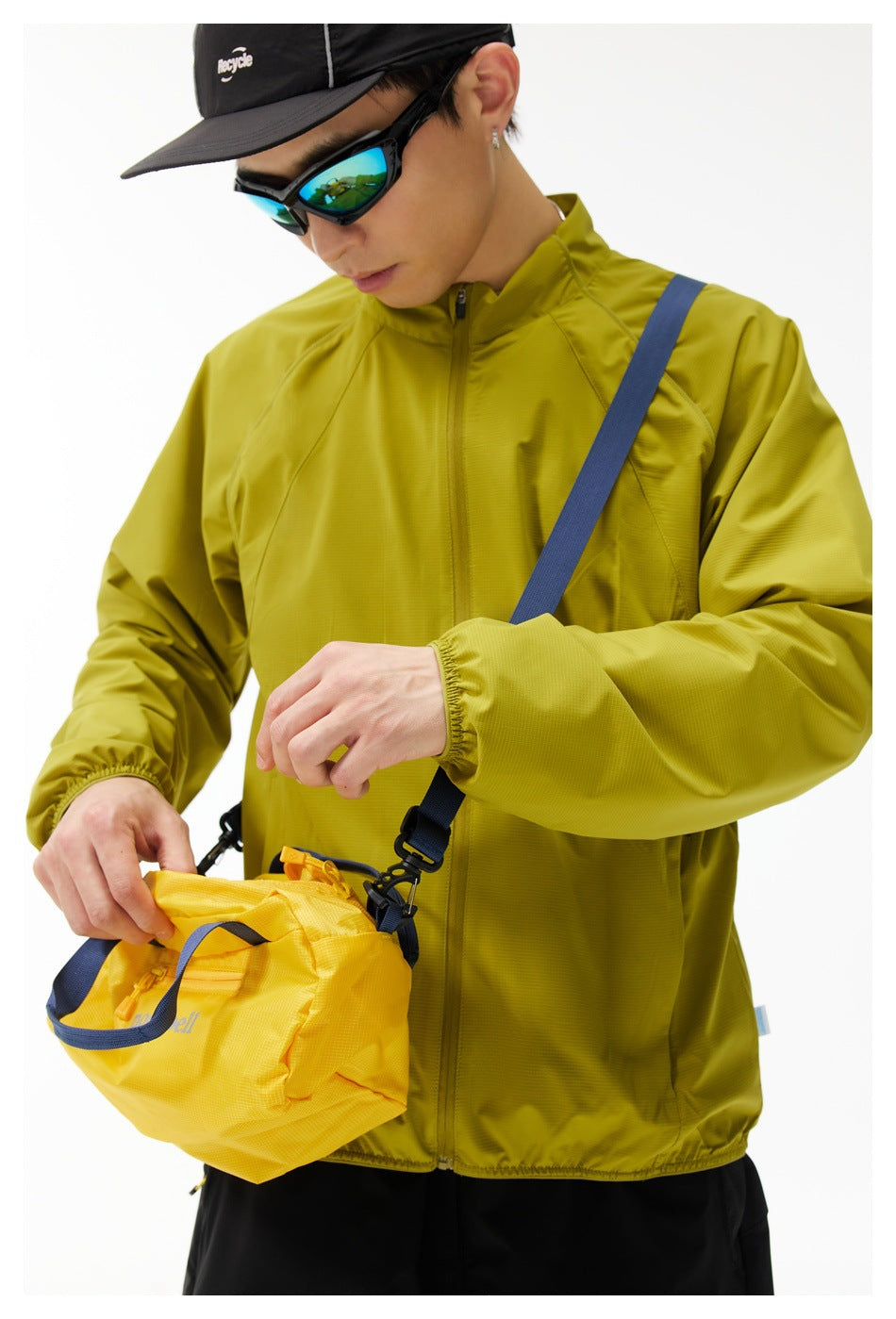 Model wearing a mustard yellow-green zip jacket with a stand collar and elastic cuffs, carrying a yellow shoulder bag.