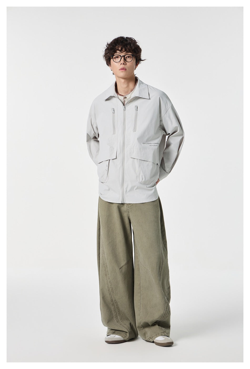 Model wearing a pale gray collared zip jacket with chest zippers and large pockets, paired with loose olive trousers.