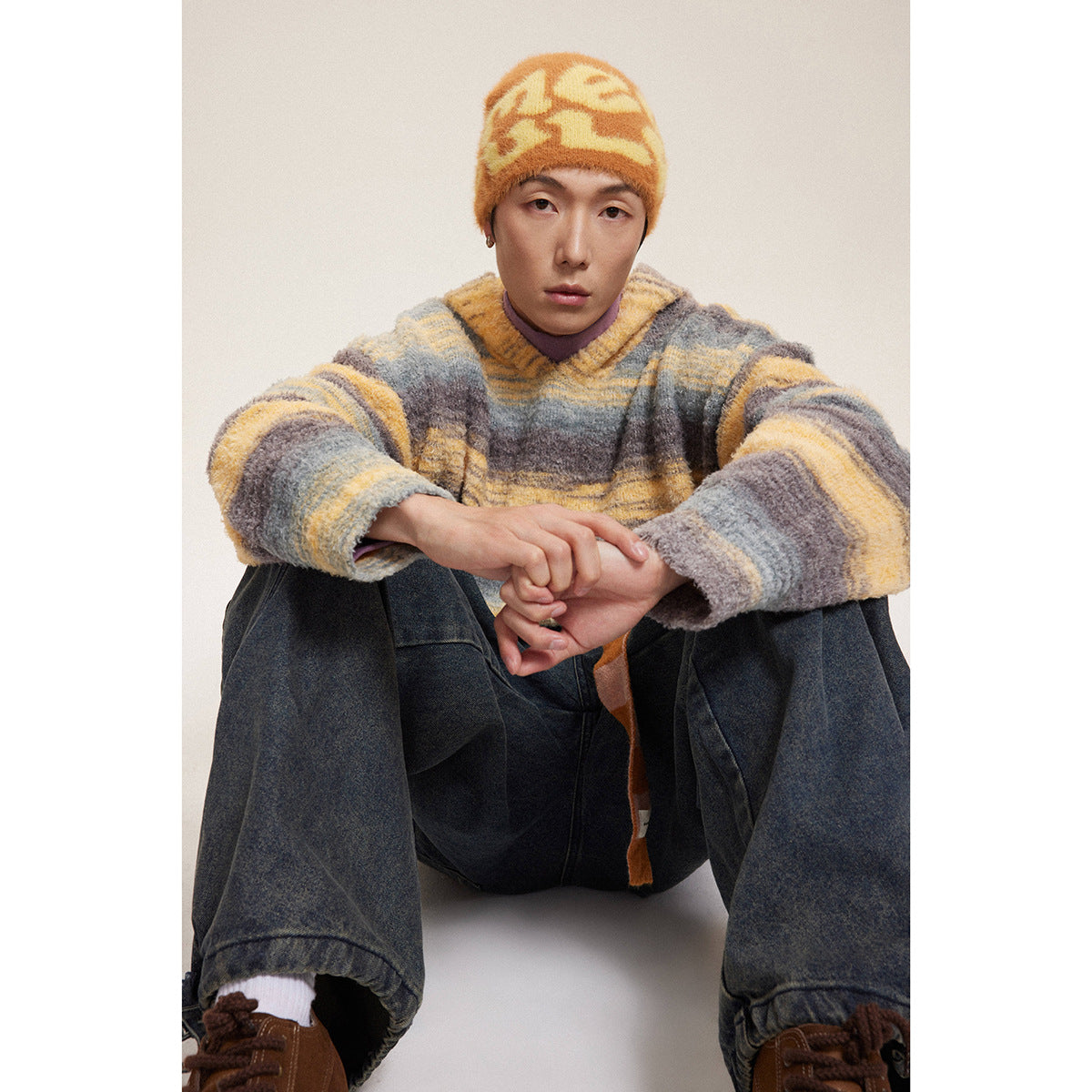 Model seated in a gray, yellow, and brown striped hooded sweater and matching orange-yellow patterned beanie.