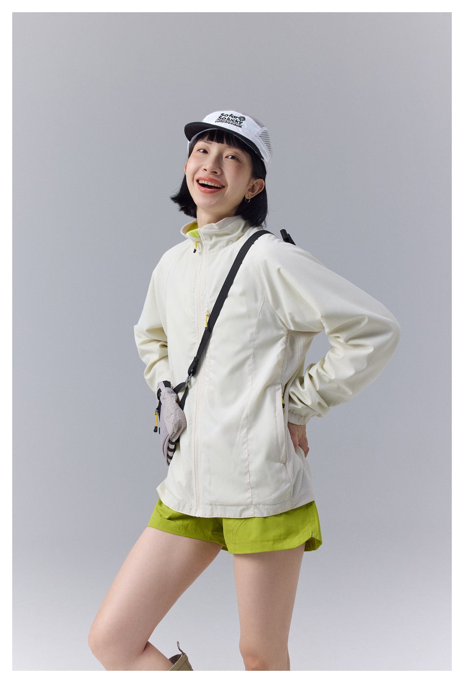Model wearing a cream zip-front jacket with lime accents and bright green shorts, carrying a small crossbody bag.