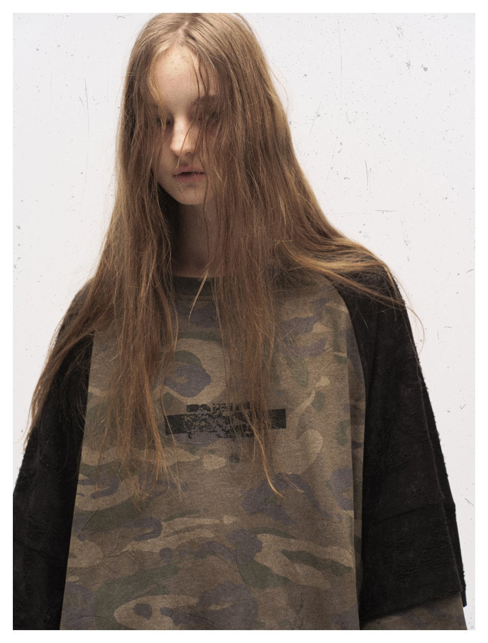 Model wearing an oversized olive camouflage top with black short sleeves and distressed black chest print, front view.