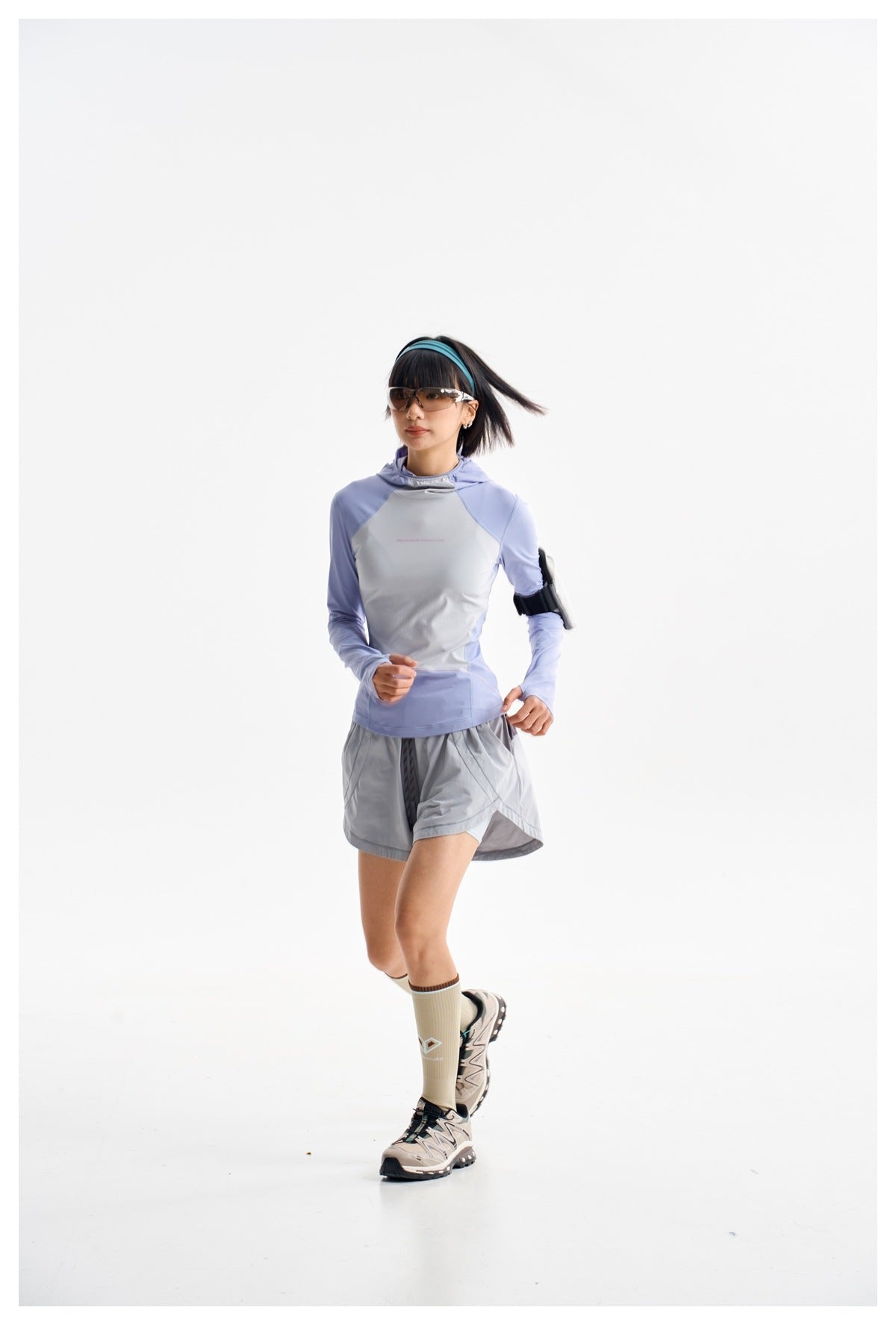 Model in a lavender and light gray hooded top and gray running shorts, captured mid-stride in sunglasses.