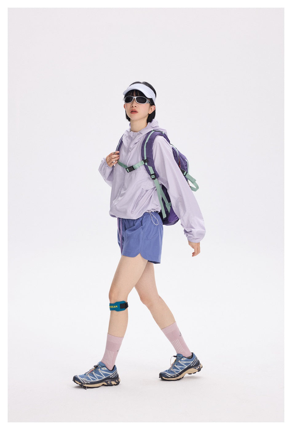Model walking in a lavender hooded zip jacket with visor, sunglasses, backpack, blue shorts and sneakers