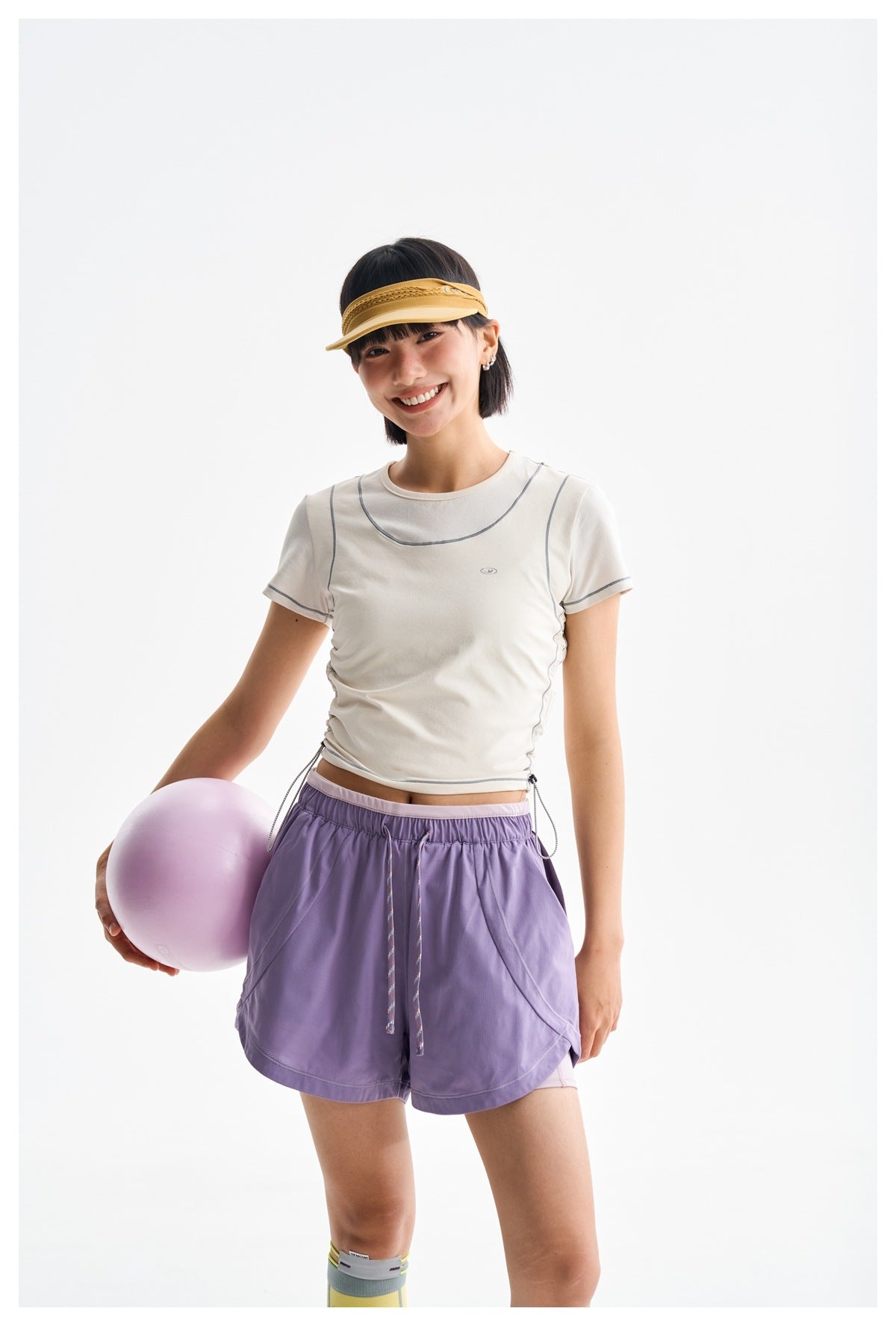 Model wearing a cream fitted tee with gray piping and lavender drawstring shorts, holding a pale purple ball.