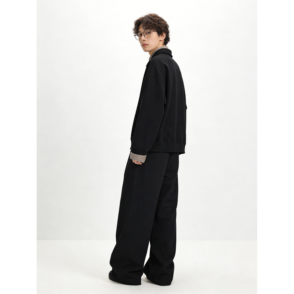 Rear three-quarter view of a model wearing a black pullover and loose black trousers with plaid cuffs.