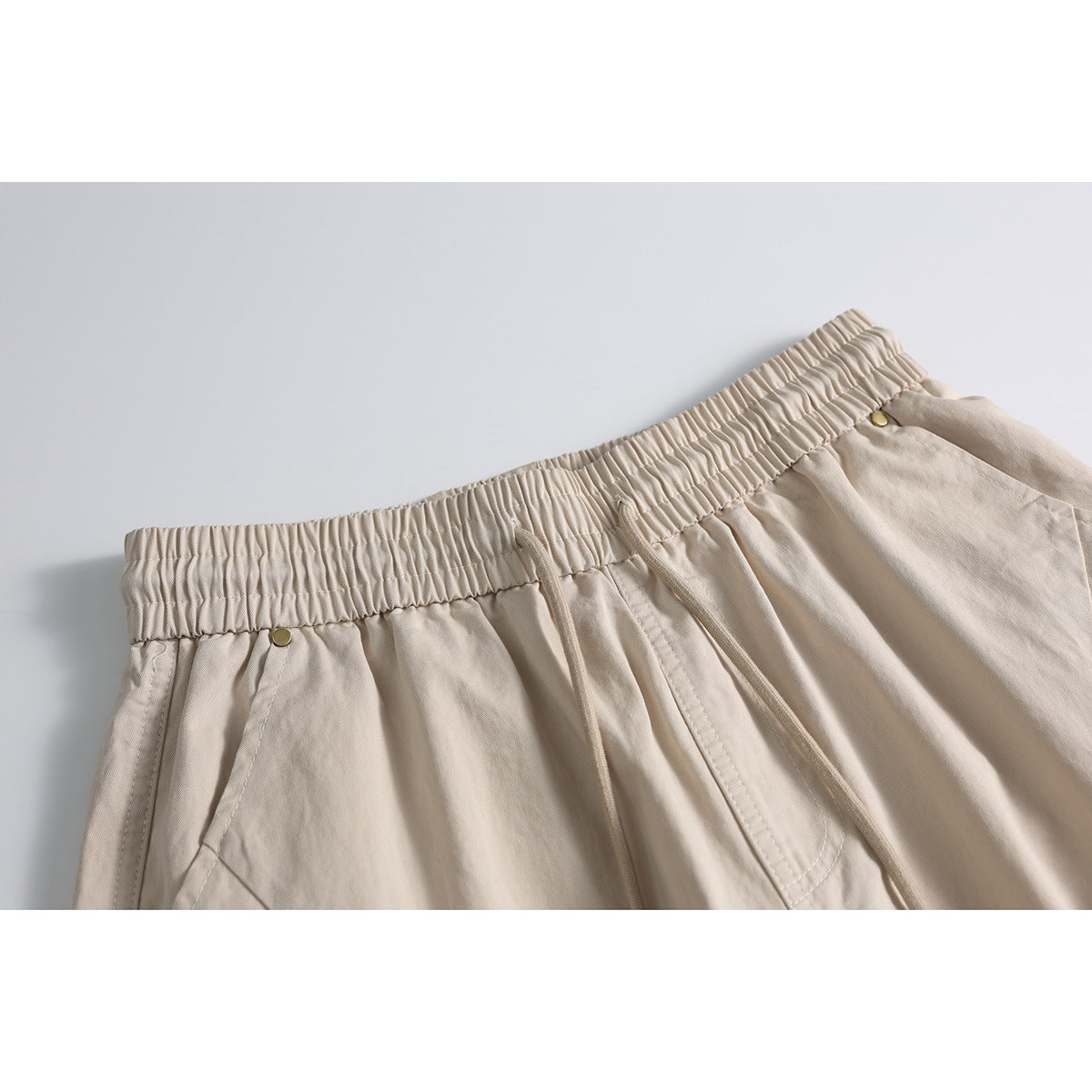 Close-up of beige shorts showing the gathered elastic waistband, drawstrings, angled pocket seams and small metal rivets.